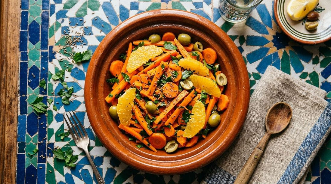 Moroccan carrot salad — sliced carrots with orange segments, green olives, parsley, and cumin in a terracotta bowl on a zellige-tiled table
